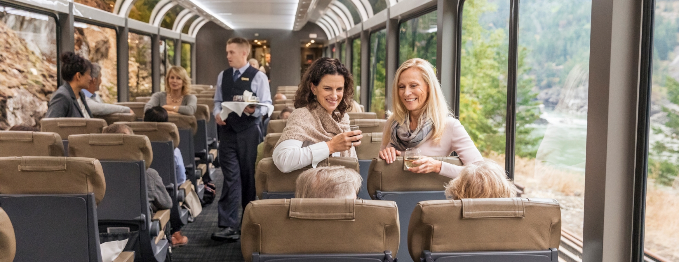 Two women onboard rocky mountaineer train in silverleaf service with wine leaning over chairs