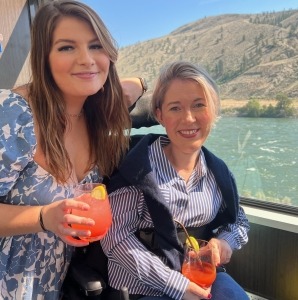 A pair of women smiling at the camera holding cocktails beside a window with a glacial green river outside while onboard the Rocky Mountaineer train.
